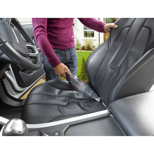Person using a BLACK+DECKER handheld vacuum cleaner to clean crumbs from the leather seat inside a car.