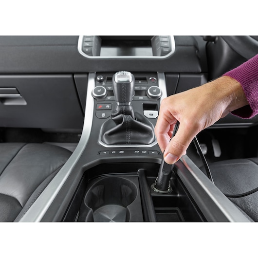 A person is plugging a device into the power socket located in the center console of a car interior. The console shows the gear shift, cup holders, and control buttons.
