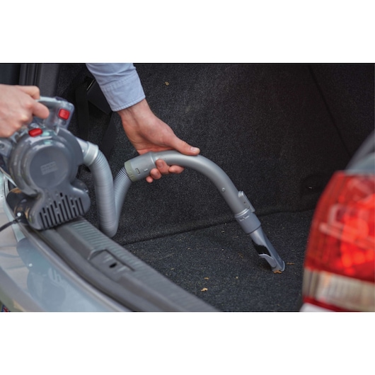 A person is using a Black+Decker handheld vacuum cleaner with a flexible hose attachment to clean the interior of a car trunk.