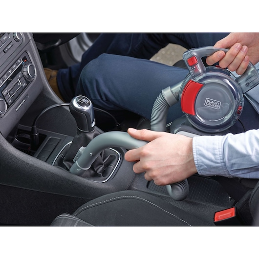A person is using a BLACK+DECKER portable vacuum cleaner, model PD1200AV, to clean the interior of a car. The vacuum is being used near the gear shift area, and the flexible hose is visible.