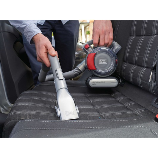 A person uses a BLACK+DECKER Dustbuster Flexi Auto handheld vacuum cleaner to clean the seat of a car. The vacuum is grey and red and has a flexible hose attachment for reaching tight spaces.