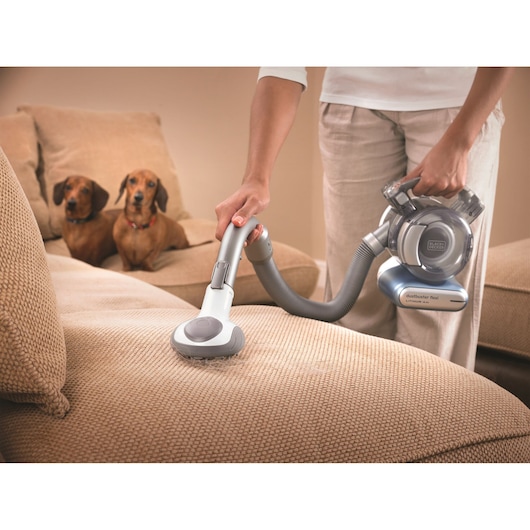 A person uses a BLACK+DECKER Dustbuster Flex handheld vacuum cleaner to clean a beige sofa. Two small dogs are sitting on the sofa in the background.
