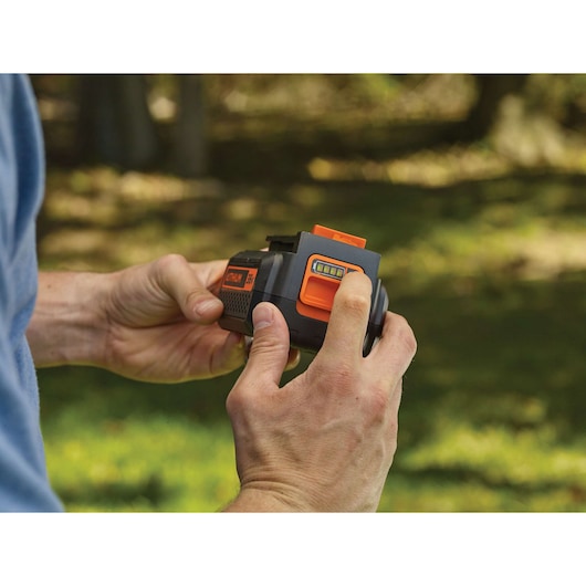 Close-up photo of hands holding BLACKDECKER STB3620L battery with orange button and charge indicator outdoors.