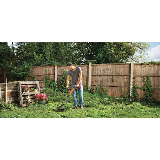 Person using BLACKDECKER STC1840PC cordless grass trimmer in a garden with wooden fence and flowers.