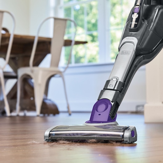 A BLACK+DECKER cordless vacuum cleaner with purple accents cleaning up pet hair from a hardwood floor in a modern dining area, with chairs and a table in the background.