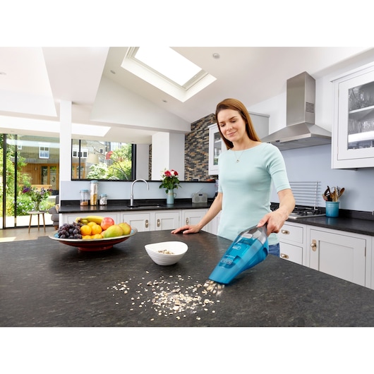BLACKDECKER WDC215WA handheld vacuum in blue used to clean oats from kitchen counter near fruit bowl.