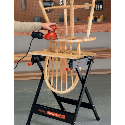 A wooden chair is being sanded using an electric sander, supported by a BLACK+DECKER Workmate 301 portable workbench in a workshop.