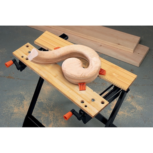 A BLACK+DECKER WM301 workbench holds a curved wooden piece securely with orange clamps. The bench has a light wooden surface and black metal legs, with wooden boards in the background.