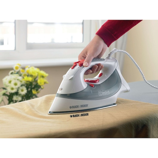 BLACKDECKER XT2030 steam iron held over beige fabric, white and grey body, red buttons, indoor setting.